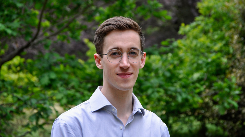 A man wearing glasses and a light blue shirt, smiling, with green brushes and leaves behind him. A man wearing glasses and a light blue shirt, smiling, with green brushes and leaves behind him.
